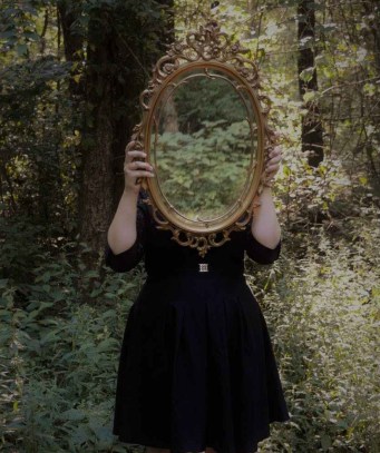 woman holding mirror against her head in the middle of forest